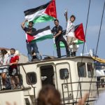 Anti-war Activists stand aboard a ship waving large Palestinian flags, engaging with a crowd onshore during a pro-Palestinian demonstration.
