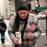 Homeless man holding U.S. flag and coffee on NYC street, symbolizing the human cost of Trump’s shutdown and neglect of vulnerable Americans.