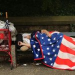 A homeless American sleeps on park bench under American flag blanket beside a cart of belongings, symbolizing poverty under Trump’s failed economic vision.