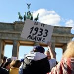 A large crowd gathers at Brandenburg Gate in Berlin, holding signs protesting Trump's National Security Strategy. The demonstration emphasizes the need to defend democratic values and transatlantic alliances against rising isolationism and extremist rhetoric.