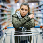 A young woman with a sad, worried expression leans on an empty shopping cart in a brightly lit grocery store aisle. Her posture conveys distress, reflecting the growing economic anxiety and cost of living challenges many Americans face under the Trump presidency.